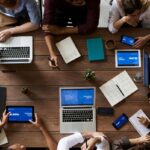Grupa osób korzystająca z przestrzeni do co-workingu. Kadr skierowany na stół, na którym leżą laptopy, kalendarze, długopisy