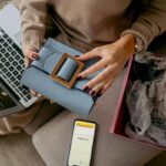 Kobieta rozpakowująca paczkę z zakupów online, trzymająca w ręce torebkę, a na kolanach laptopa.