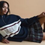 Black Friday 2024 w sieci sklepów Inditex. Stradivarius to marka, która zaoferuje klientom 50% rabaty na Black Friday. Na zdjęciu kampania AW2024 Stradivarius - modelka z długimi ciemnymi włosami pozuje leżąc na beżowym tle. Na na sobie bluzę w stylo collage z zamkiem pod szyją i białą wstawką oraz napisem oraz krótką plisowaną spódniczkę w kratę w stylu preppy.