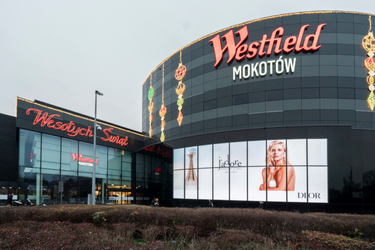 Zdjęcie przedstawia fasadę centrum handlowego Westfield Mokotów w Warszawie, ozdobioną świątecznym napisem „Wesołych Świąt” oraz reklamą perfum Dior. W tle pojawia się slogan „napad w galerii mokotów”, który przykuwa uwagę i stanowi nawiązanie do potencjalnego zdarzenia w tej lokalizacji.