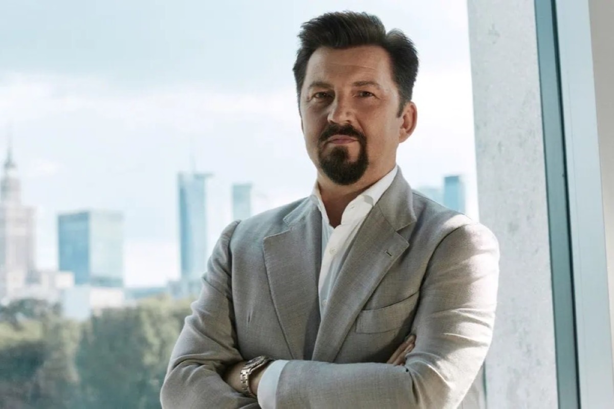 Na zdjęciu znajduje się Dariusz Miłek, prezes CCC. Mężczyzna ubrany w białą koszulę oraz beżową marynarkę, posiadający ciemne włosy i przystrzyżoną brodę.