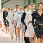 Chanel zwolnienia. Na zdjęciu Chanel pokaz mody, na którym modelki idą w rzędzie po wybiegu, prezentując eleganckie i wyrafinowane stroje. Kreacje wyróżniają się dopasowanymi krojami, wyrazistymi sylwetkami i luksusowymi tkaninami.