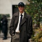 Na zdjęciu młody mężczyzna pozuje w eleganckim, jesienno-zimowym zestawie o klasycznym charakterze. Ma na sobie długi, ciemny płaszcz o dwurzędowym kroju, białą koszulę, czarny krawat oraz spodnie w odcieniu oliwkowej zieleni. Jego stylizację dopełnia czarna czapka z daszkiem z logotypem YSL, która nadaje całości nowoczesnego, casualowego akcentu. Mężczyzna stoi w otoczeniu zieleni i architektury w tle, co sugeruje miejskie, stylowe otoczenie. Jego nonszalancka poza z rękami w kieszeniach podkreśla swobodę i pewność siebie.