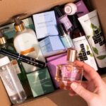 Oriflame zamyka fabrykę w Polsce. Na zdjęciu karton z kosmetykami Oriflame. Na pierwszym planie widać dłoń kobiecą trzymającą flakon perfum.