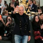 Ralph Lauren wyróżnienie – Ralph Lauren, światowej sławy projektant mody, machający do zgromadzonej publiczności podczas uroczystego wydarzenia. W tle widać ludzi robiących zdjęcia i bijących brawo, doceniając jego osiągnięcia i wkład w modę. Elegancka atmosfera z detalami w złocie i wyrafinowanymi strojami gości podkreśla prestiż okazji.