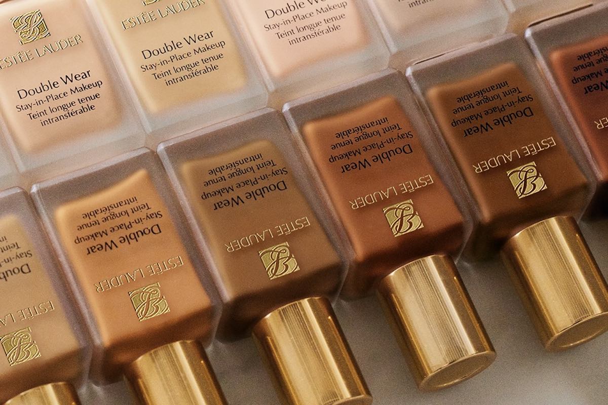 Estee Lauder szykuje masowe zwolnienia. Rząd prostokątnych butelek podkładu Estee Lauder Double Wear w różnych odcieniach (od jasnych po ciemniejsze), z charakterystycznymi złotymi nakrętkami i logo marki na froncie.