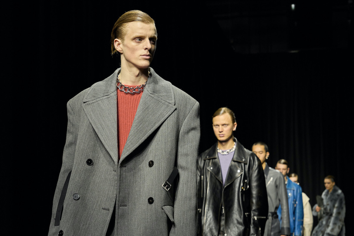Model na czele pokazu mody prezentuje oversize'owy, szary płaszcz o szerokich ramionach, połączony z czerwonym swetrem i masywnym, srebrnym łańcuchem na szyi. Za nim w tle widać innych modeli w stylizacjach z kolekcji Jil Sander, ubranych w skórzane kurtki, futra oraz elementy w odcieniach czerni, niebieskiego i szarości.