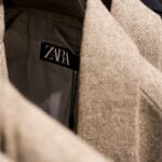 Zbliżenie na metkę z logo ZARA wszytą w wewnętrzną część jasnoszarej marynarki z delikatnym wzorem jodełki. Ubrania wiszą na wieszakach w sklepie odzieżowym, częściowo widoczne są także inne marynarki w tle.