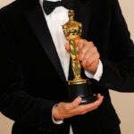 Oscarowe torby prezentowe 2025. Zdjęcie przedstawia zwycięzcę Oscarów w 2025 roku. Fokus na statuetkę, widać też męską sylwetkę w garniturze z białą koszulą i muchą.