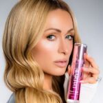 Paris Hilton reklamująca swoją nową markę kosmetyków skincare - Parívie, trzymająca w dłoni buteleczkę z produktem.